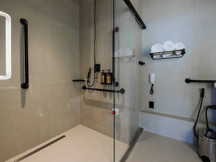 A modern bathroom with a glass shower enclosure, a towel rack with folded towels, a soap dispenser, and a handheld showerhead.