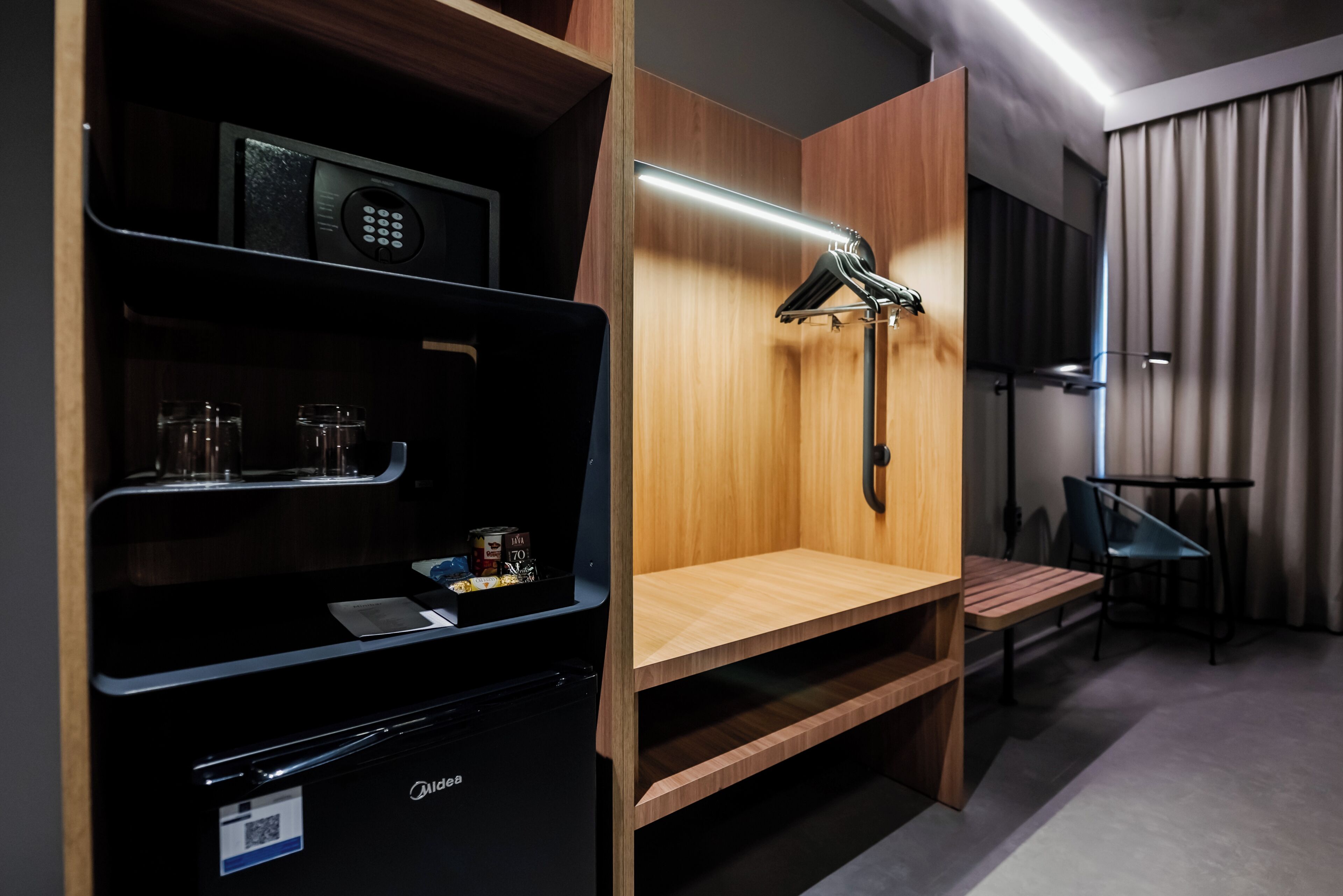 A modern hotel room with a minibar, a wooden shelf, a hanging towel rack, and a small table with chairs.
