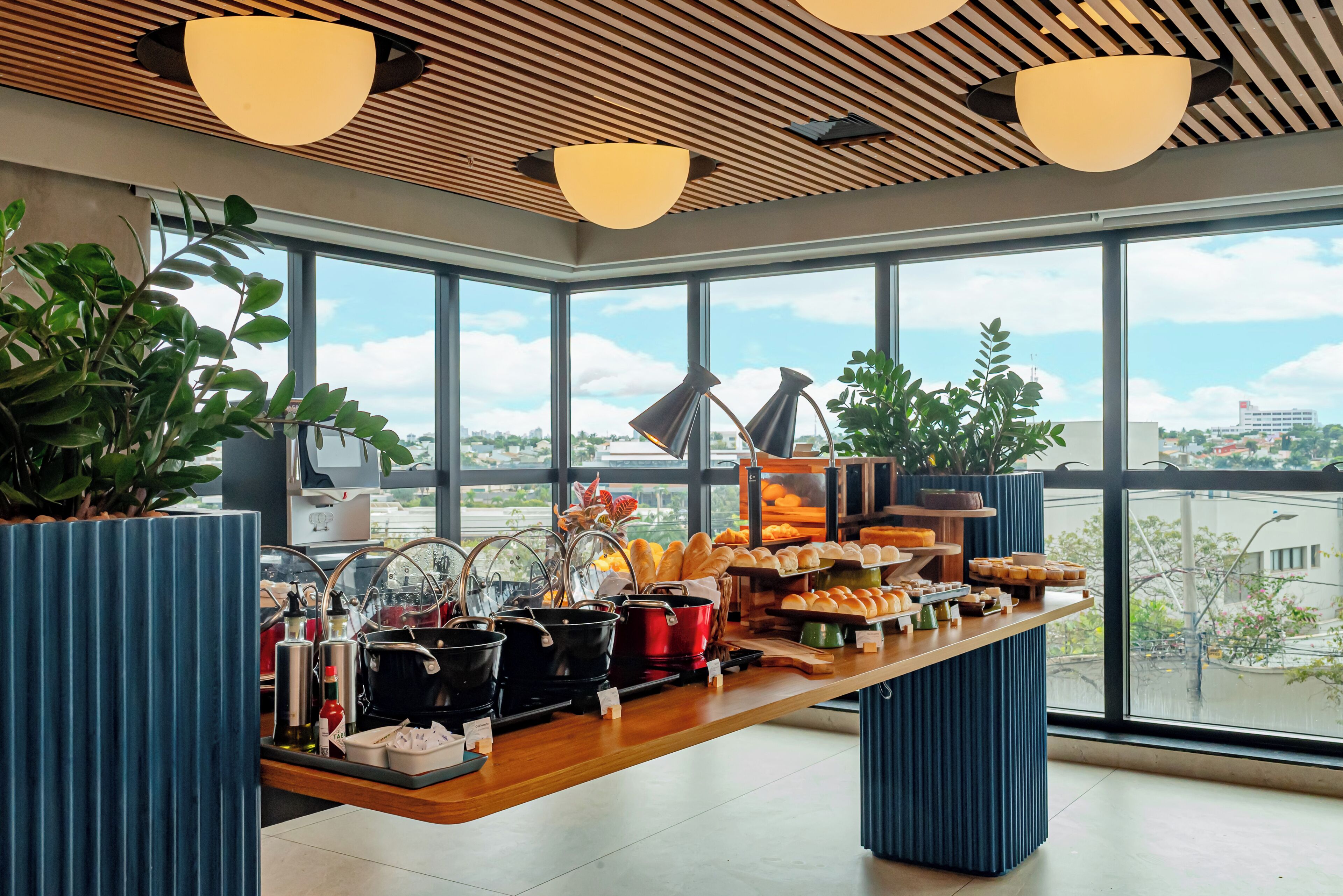 Una cocina moderna con encimera de madera que exhibe diversos alimentos y utensilios, amplias ventanas con vista a la ciudad y un techo con listones de madera y luces colgantes.