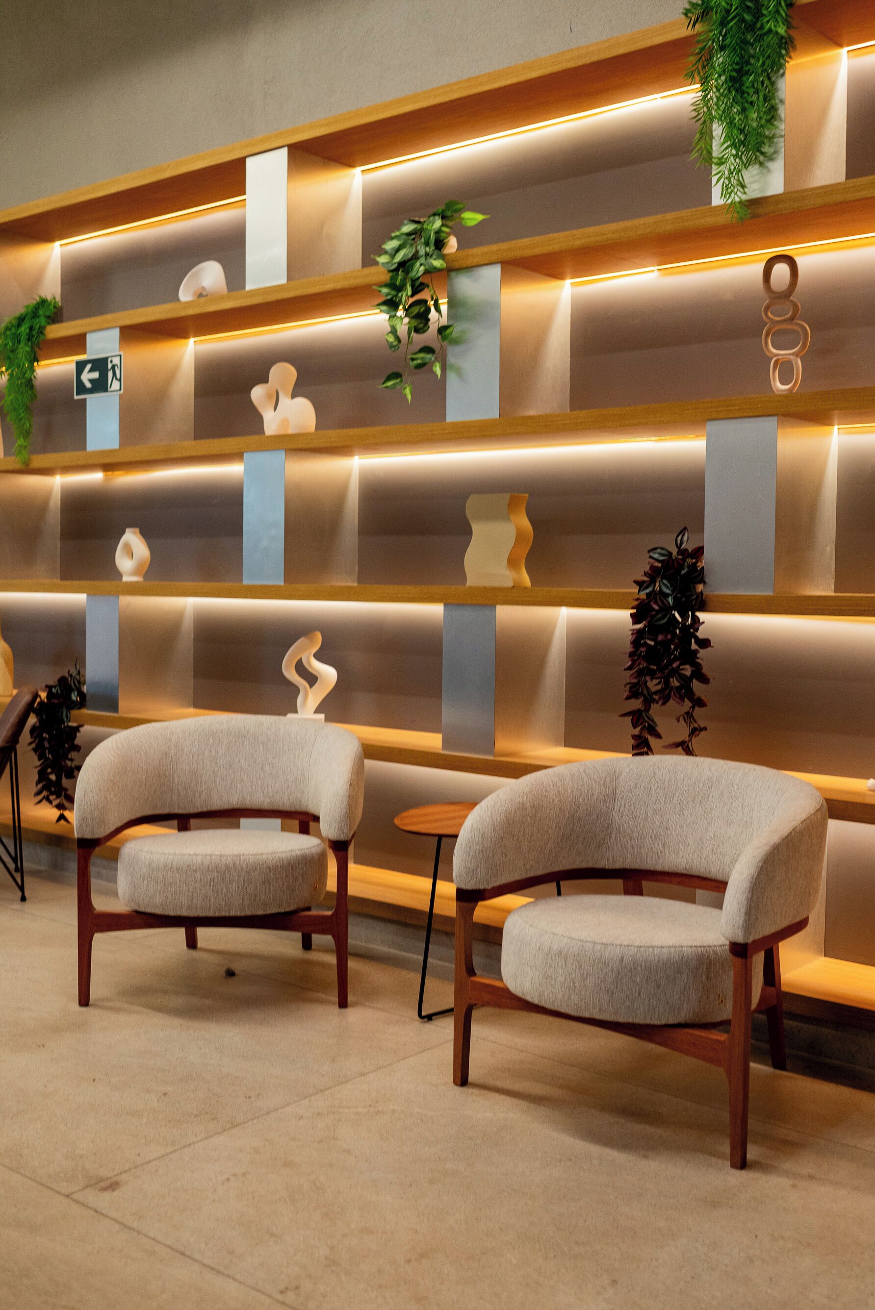 Uma área de espera moderna com duas poltronas estofadas, uma prateleira fixa com objetos decorativos e uma placa de sinalização.