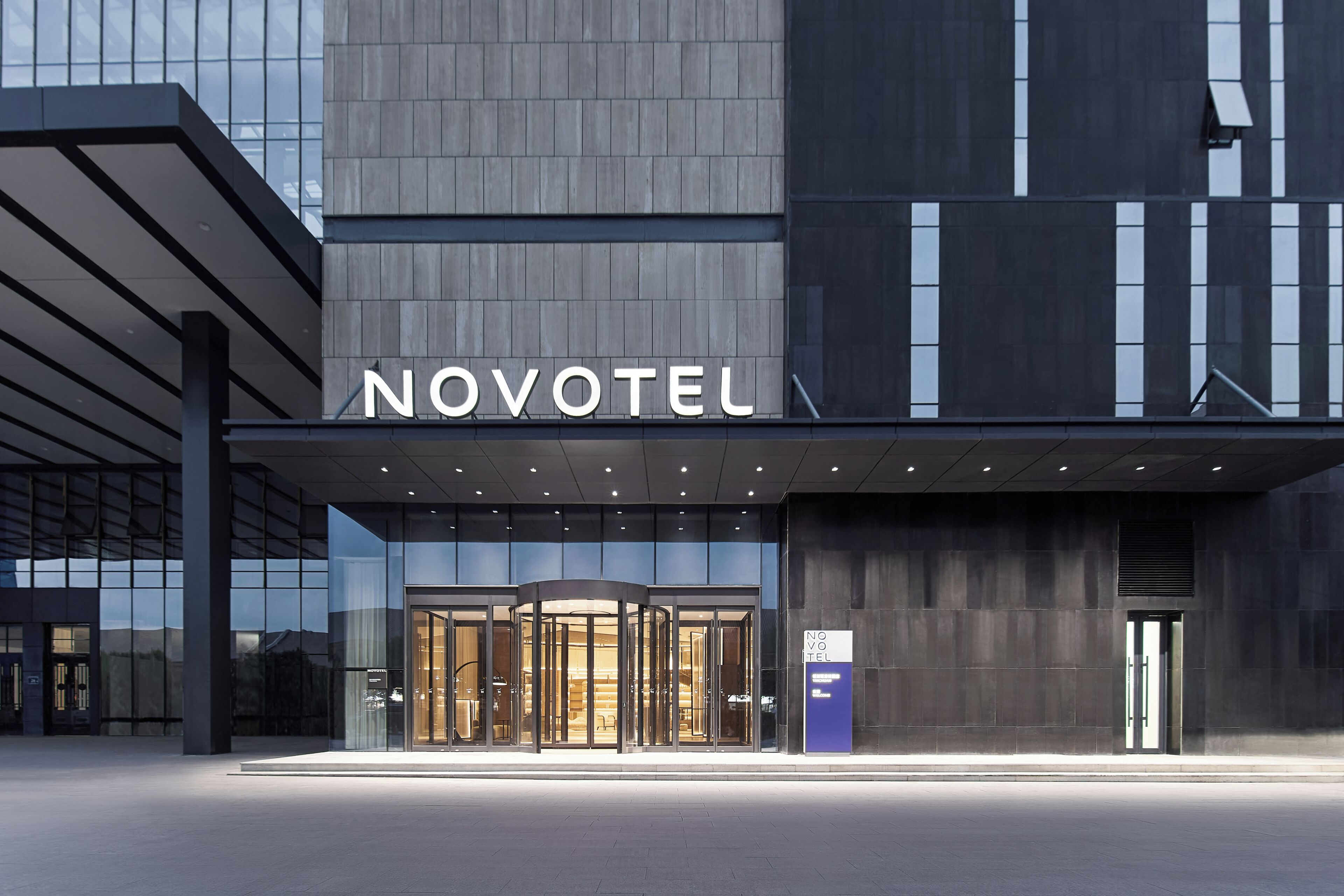 En moderne hotelindgang med en dreje-/svingdør, en glasfacade og et skilt med teksten 'NOVOTEL'.