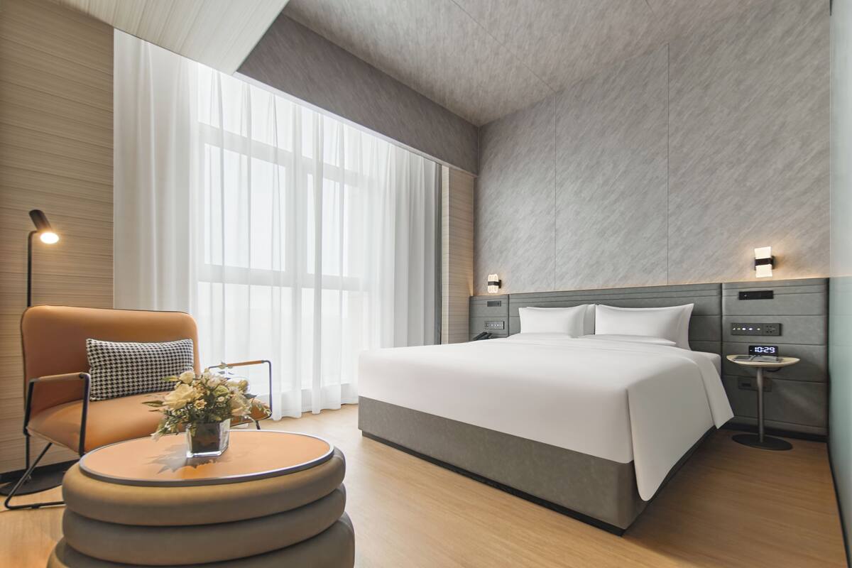 Une chambre d’hôtel moderne avec un grand lit, une table basse ronde ornée de fleurs, un canapé avec un coussin à carreaux et une table de chevet avec une horloge.