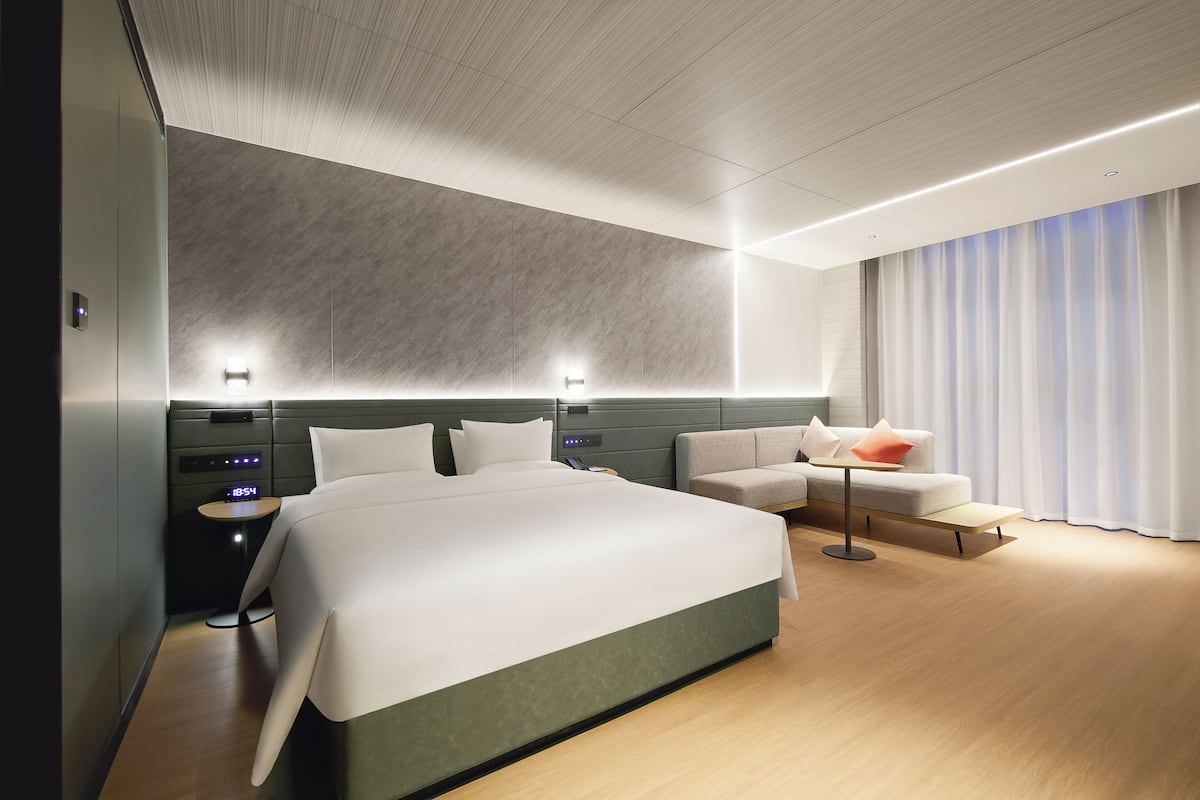 Une chambre d’hôtel moderne, dotée d’un grand lit, d’un canapé, d’une petite table et d’une table de chevet avec une horloge numérique.