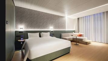 Una habitación de hotel moderna con una cama grande, un sofá, una mesita y una mesita de noche con un reloj digital.