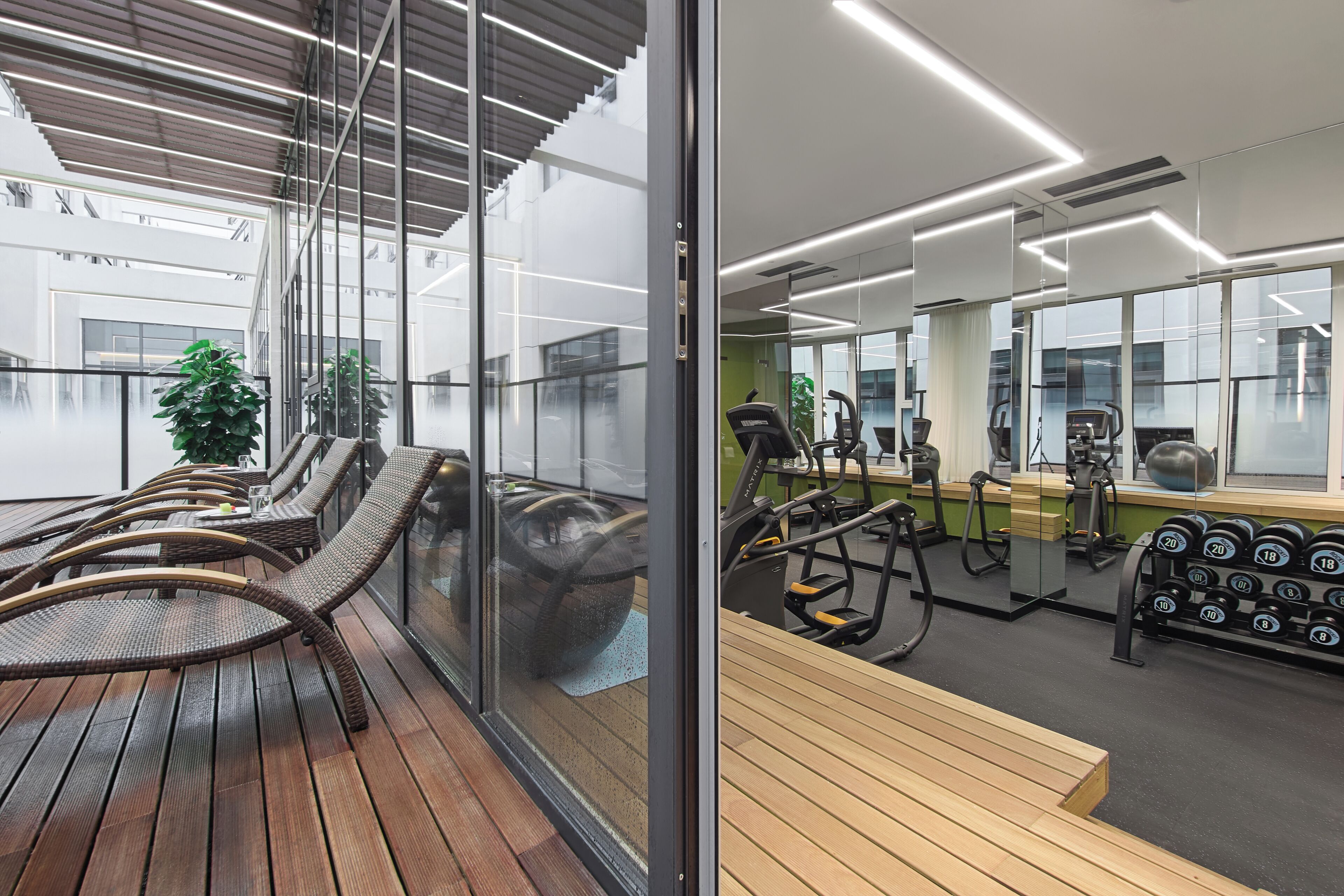 A modern gym with exercise equipment, a wooden bench, and a glass wall overlooking a balcony with wicker chairs.