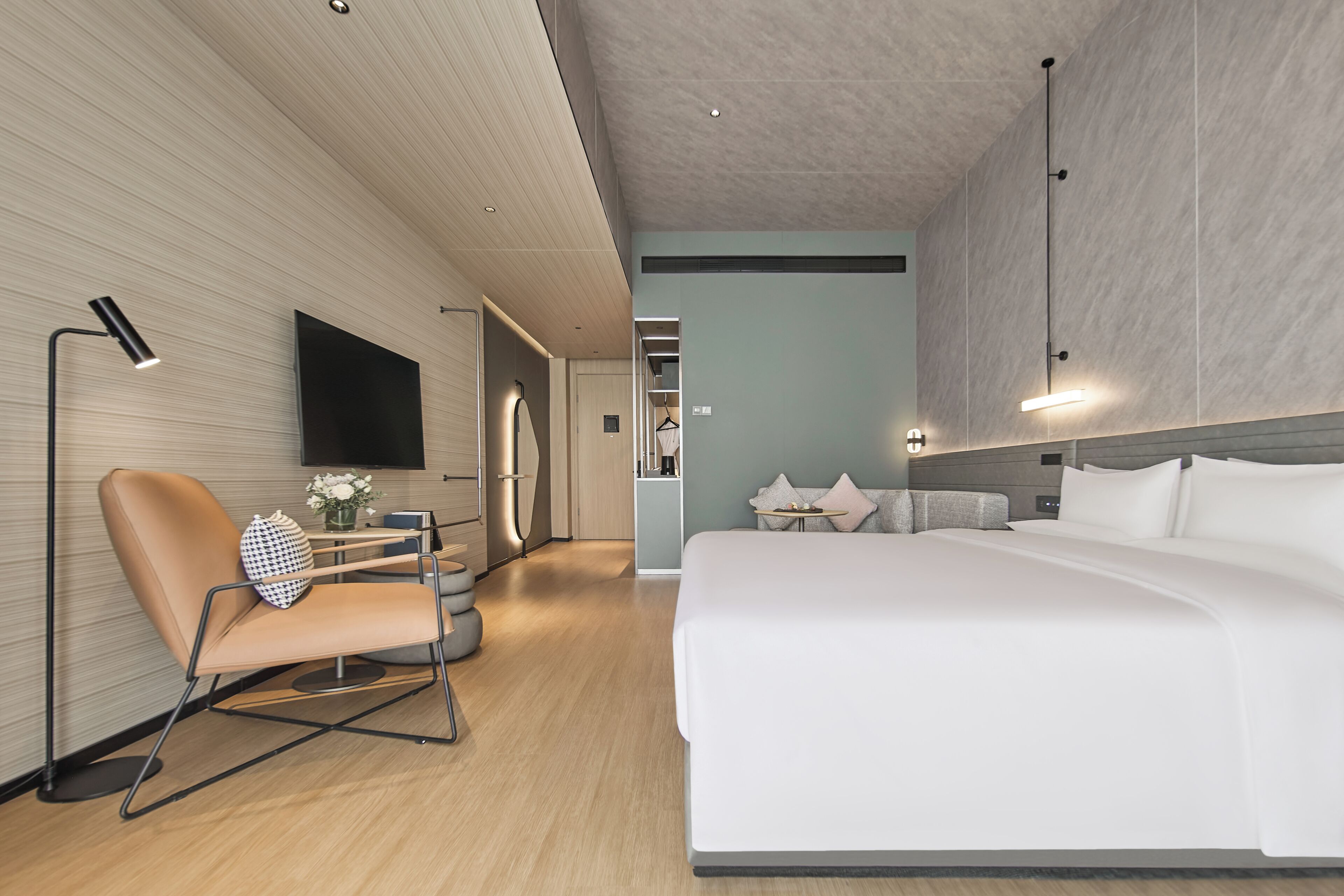A modern hotel room with a bed, a seating area with a chair, a flat-screen TV, and a hallway leading to a bathroom.