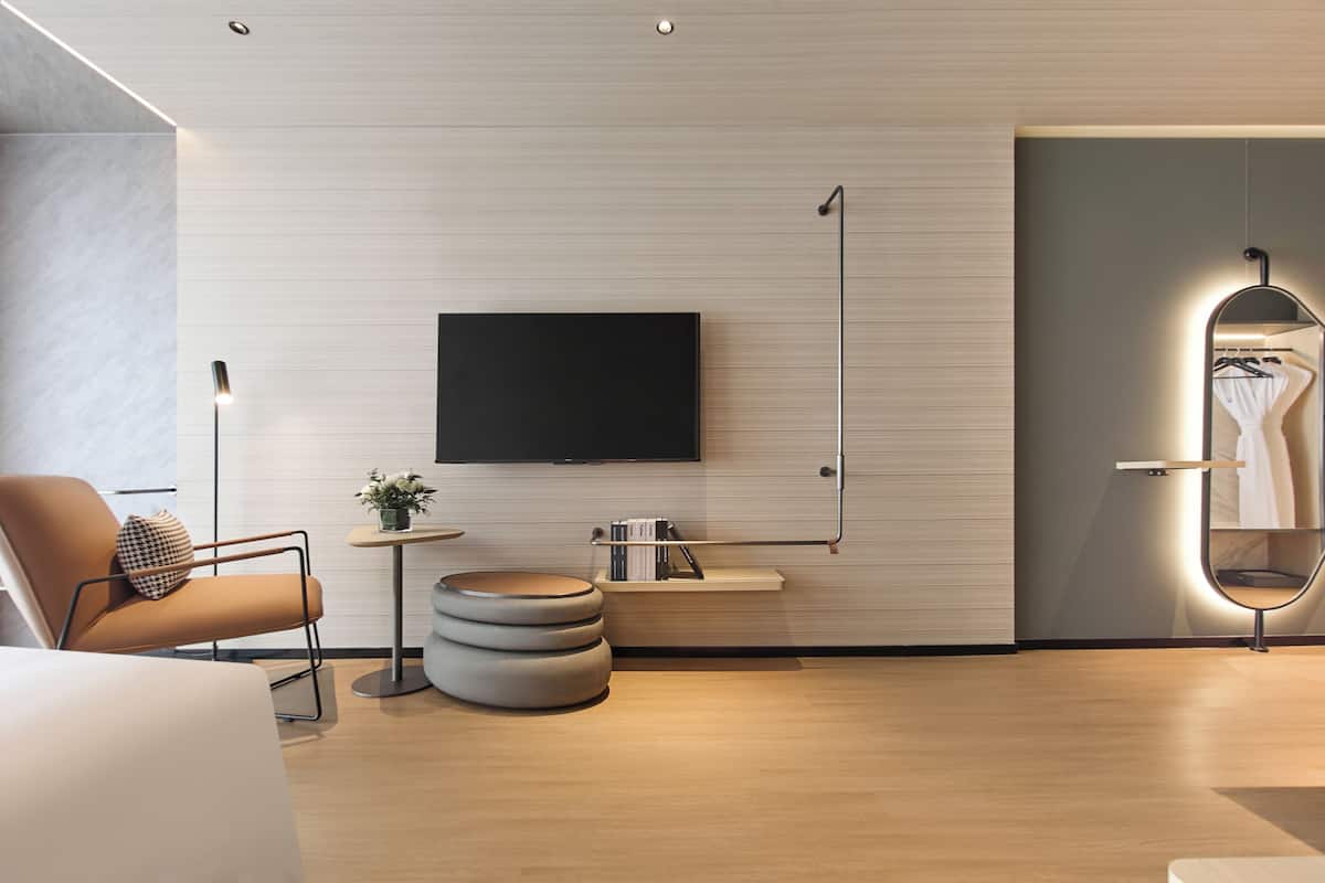 Une chambre d’hôtel moderne équipée d’un téléviseur à écran plat, d’un pouf rond, d’une petite table d’appoint et d’un miroir avec éclairage intégré.