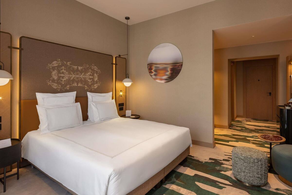 A hotel room with a large bed, a headboard with a decorative panel, a round wall mirror, a small side table, and a patterned carpet.