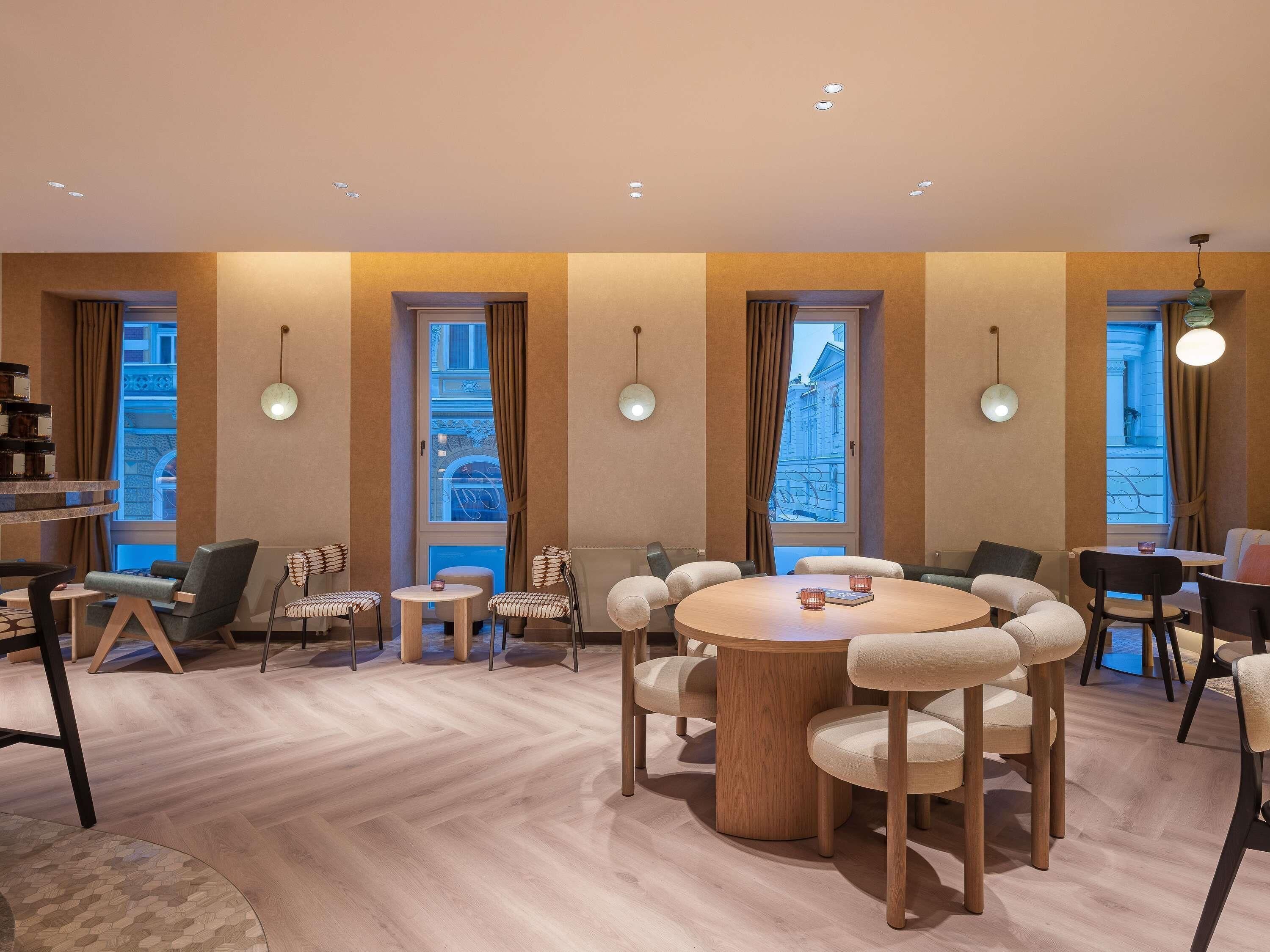 A modern hotel room with a round wooden table, multiple chairs, a small side table, and large windows with curtains.