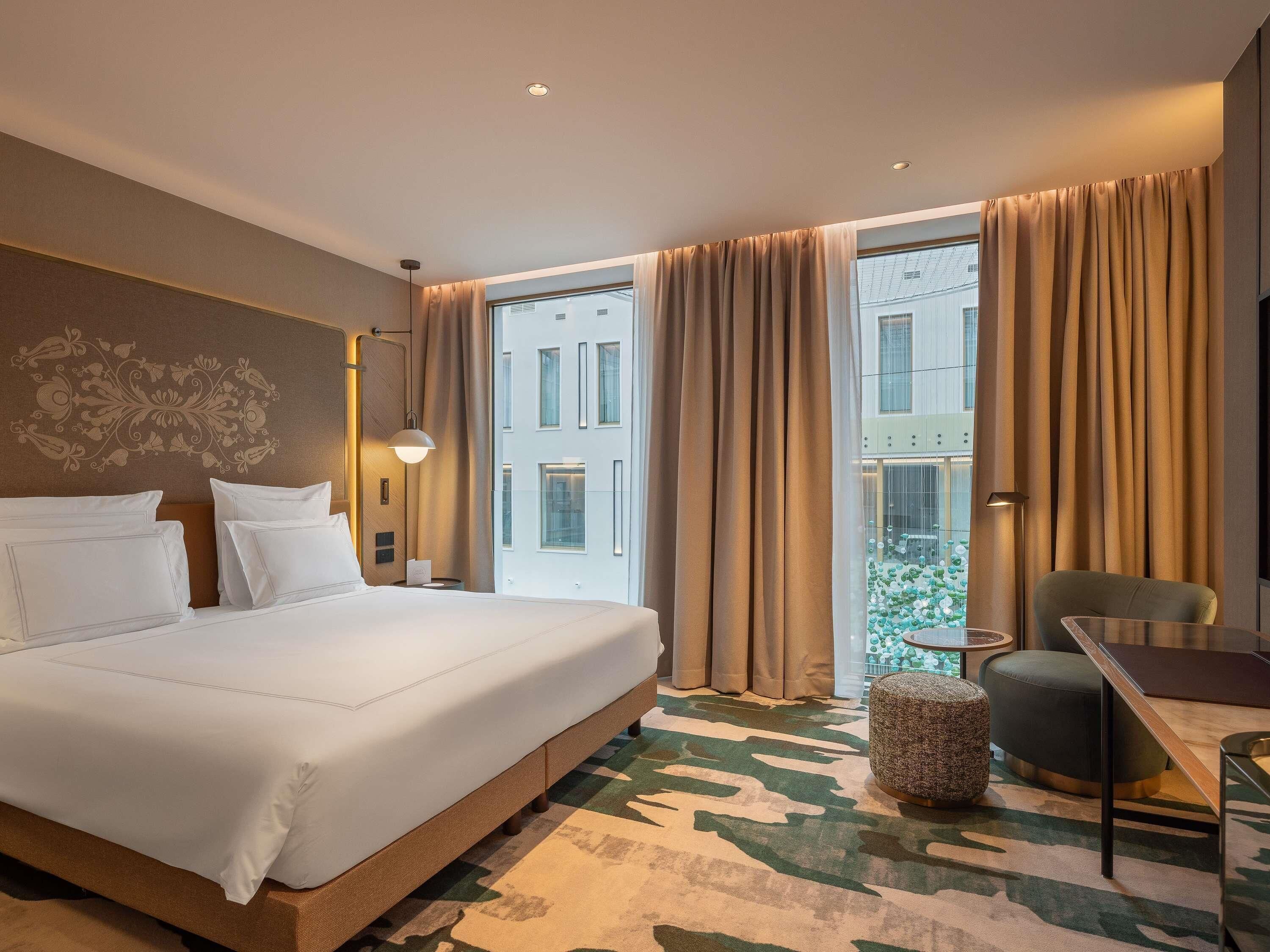 A hotel room with a large bed, a desk, a chair, a lamp, and a view of buildings through the window.