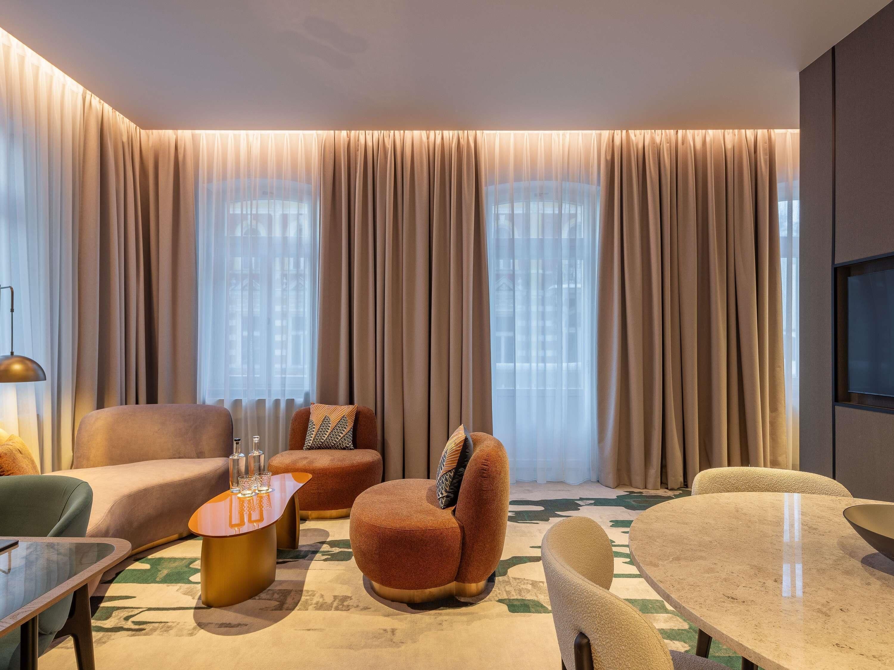 Ein modernes Wohnzimmer mit Sofa, Hocker und rundem Tisch, großen Fenstern mit Vorhängen sowie einem an der Wand befestigten Flachbildfernseher.