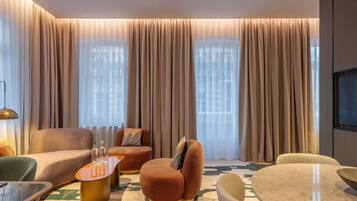 Ein modernes Wohnzimmer mit Sofa, Hocker und rundem Tisch, großen Fenstern mit Vorhängen sowie einem an der Wand befestigten Flachbildfernseher.