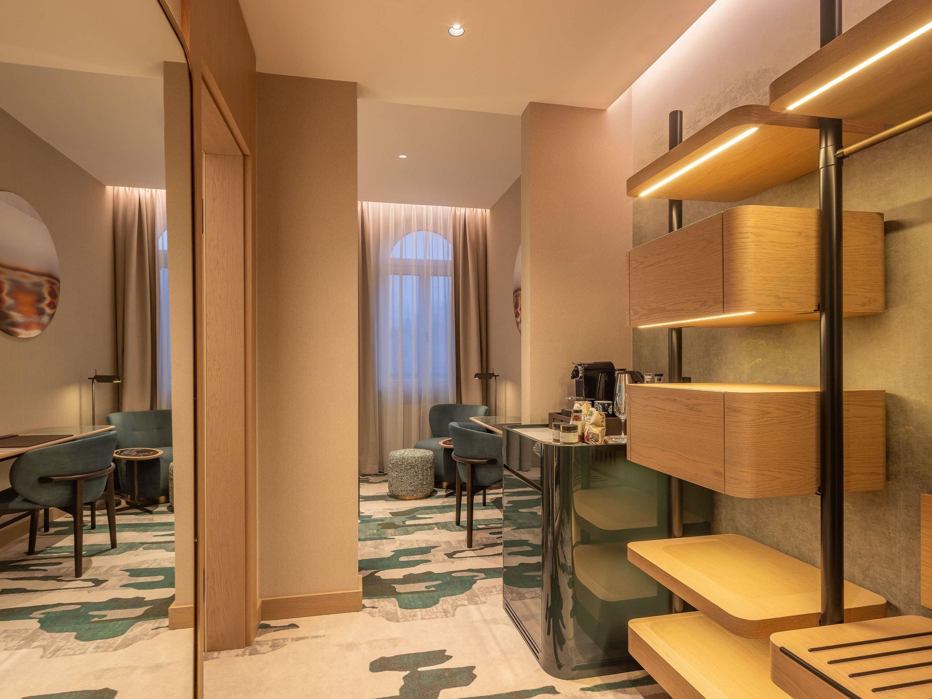 A modern hotel room with a seating area, a coffee station, and a large window with curtains.