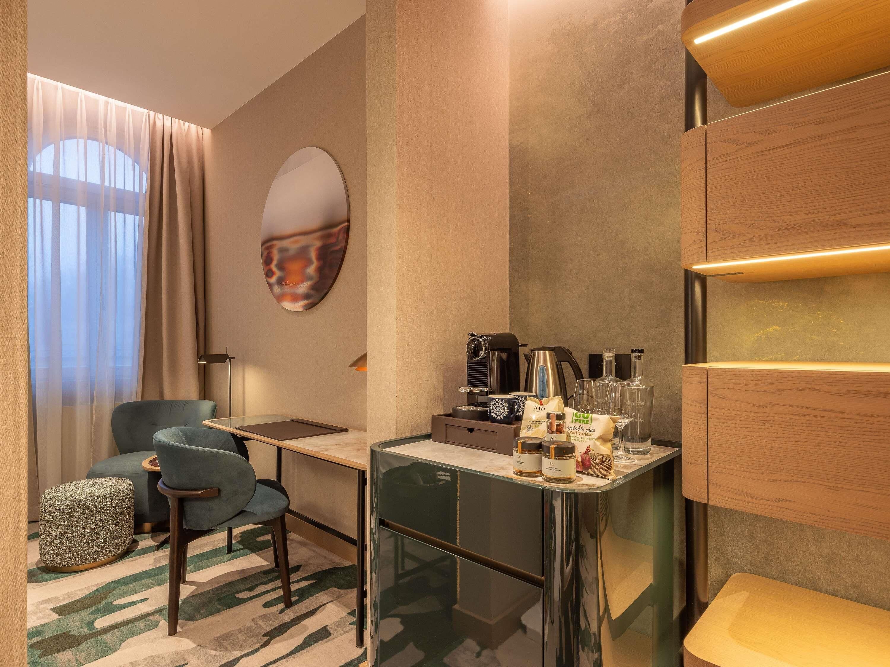 Quarto de hotel moderno com escrivaninha de vidro, sofá, poltrona, tapete estampado e um espelho grande.