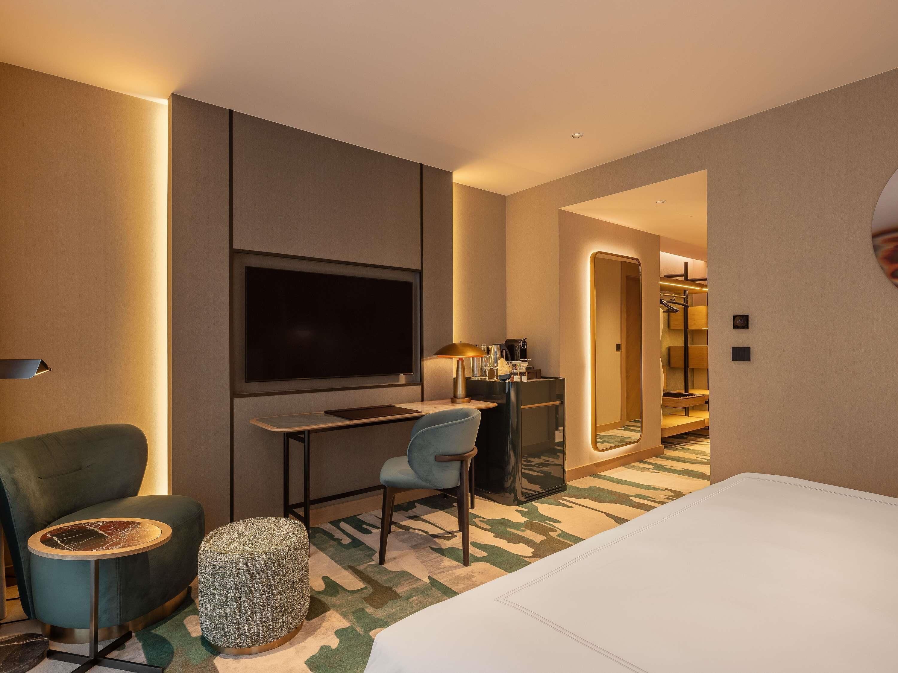 Habitación de hotel con cama, escritorio con televisor de pantalla plana, silla, reposapiés redondo y espejo.