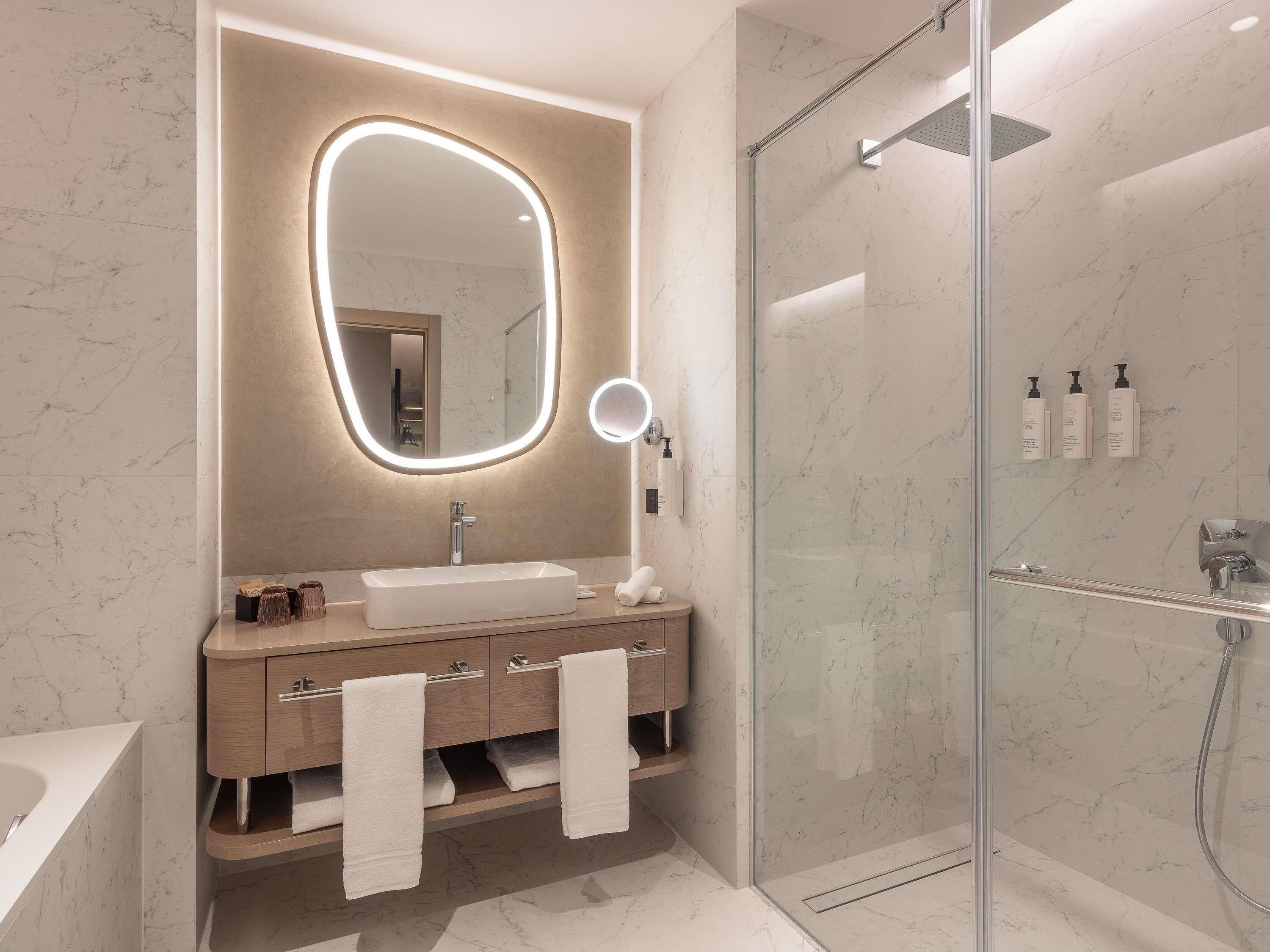 A modern bathroom with a large oval mirror, a glass-enclosed shower, and a vanity with a sink and towel rack.