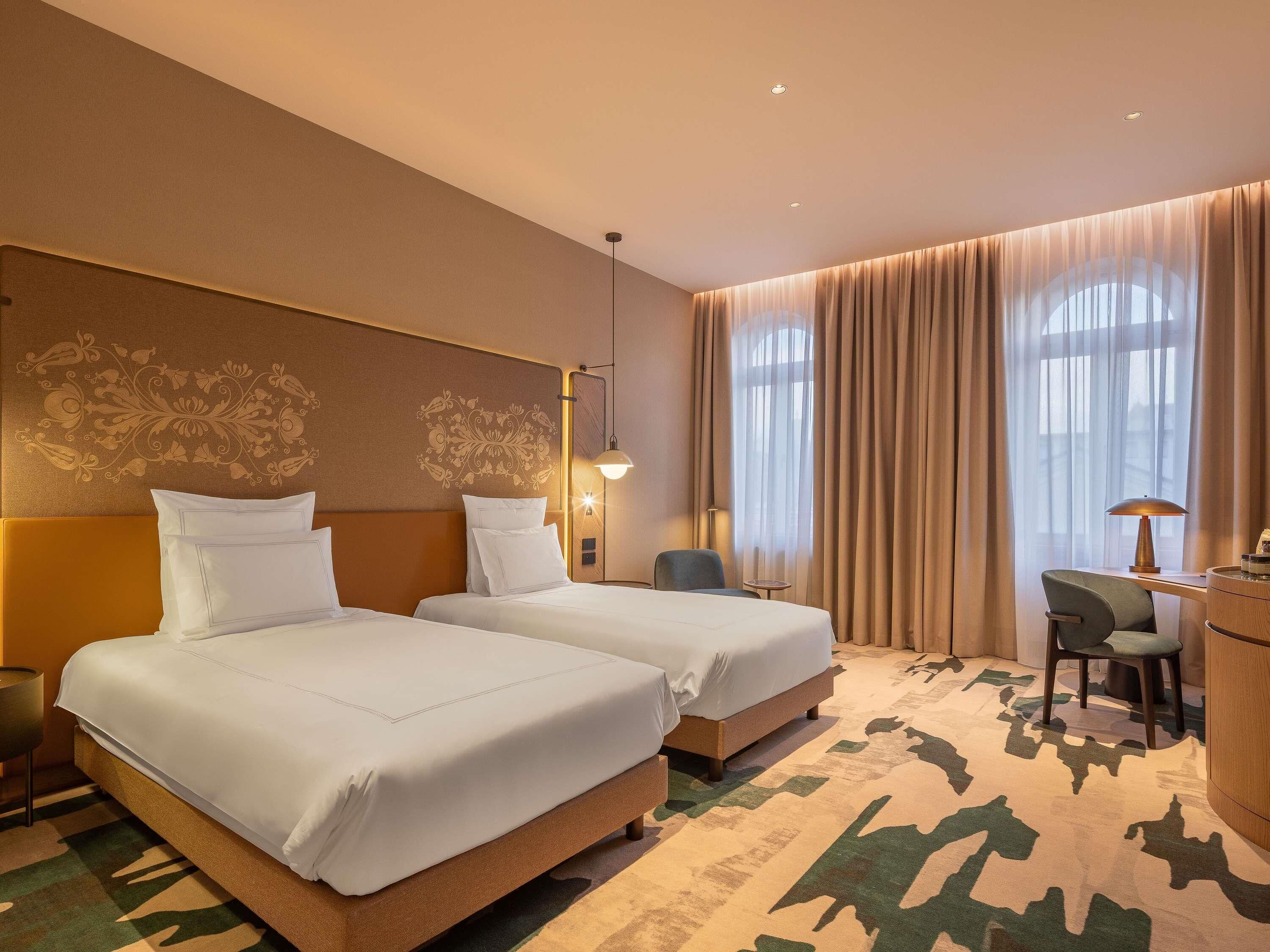 A hotel room with two beds, a desk, a chair, a lamp, and a large window with curtains.