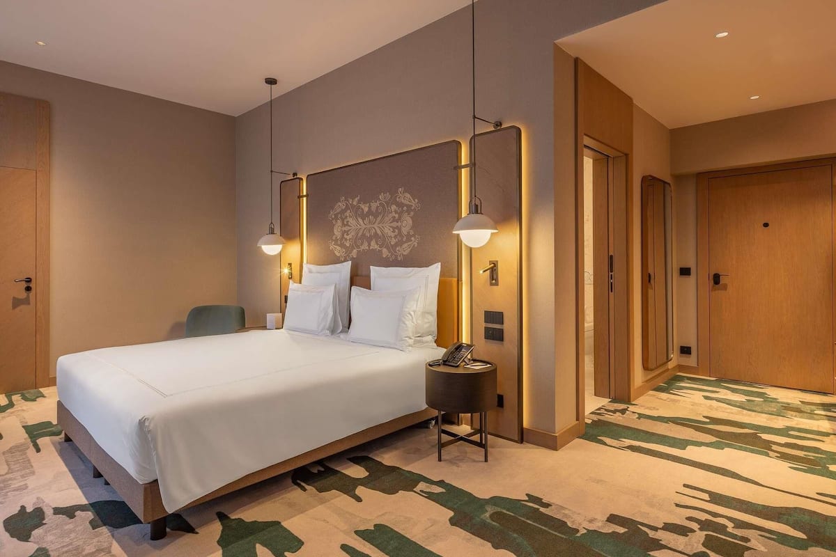 A hotel room with a large bed, two bedside lamps, a small round table with a phone, and a patterned carpet.