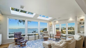 Ein geräumiges Wohnzimmer mit großen Fenstern, das Blick auf das Meer und die Berge bietet, einem blau gemusterten Teppich und verschiedenen Sitzgelegenheiten, darunter ein violettes Sessel und beigefarbene Sofas.