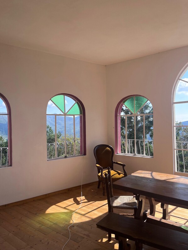 A room with wooden flooring, a wooden table, a chair, and three arched windows with views of greenery and mountains.