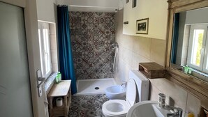 Een badkamer met een badkuip, twee toiletten, een wastafel, een douche, vloertegels met een patroon, een raam met gordijn en een spiegel.
