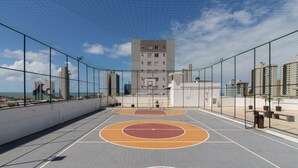Een buitenbasketbalveld met een net omheining, een basketbalring en een uitzicht op een hoog gebouw op de achtergrond.