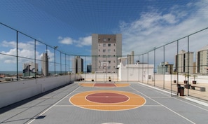 Ein Basketballplatz im Freien mit Netz umzäunt, einem Basketballkorb und im Hintergrund ein hoher Gebäudebau.