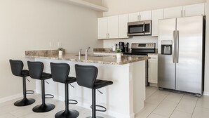 A modern kitchen with white cabinets, a stainless steel refrigerator, a microwave, and a granite countertop. There are four black bar stools at the island. - Spacious 3-Bedroom Getaway — Perfect for Families or Groups!” (Cape Coral)