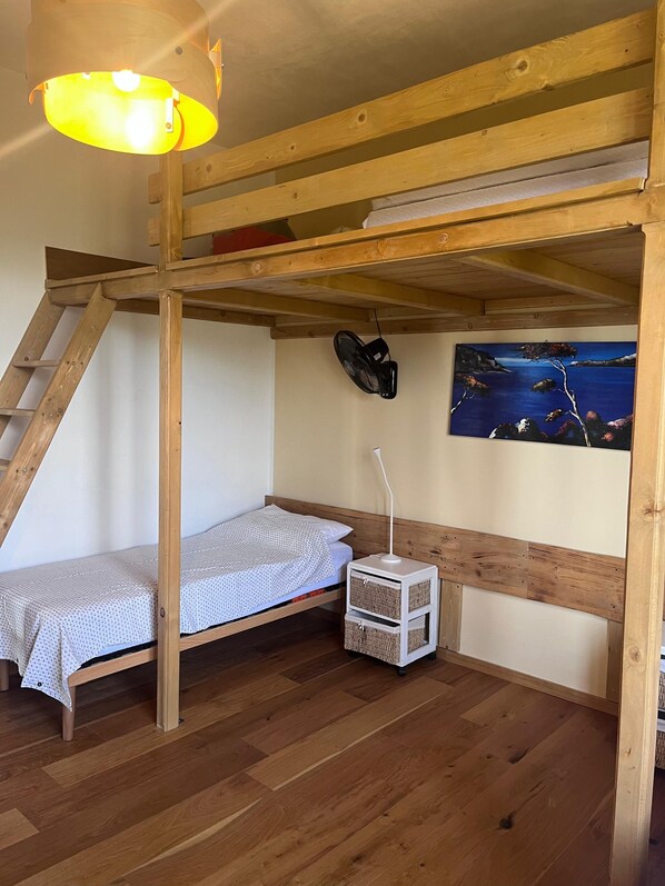 A bunk bed room with a ladder, a small white nightstand, a wall-mounted fan, and a painting of a coastal scene.