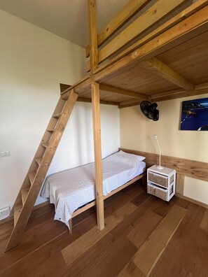 A loft-style bedroom with a wooden beam ceiling, a single bed, a small bedside table, and a wall-mounted fan.