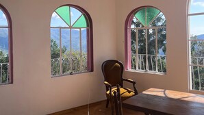 Habitación con piso de madera, una mesa y dos sillas. Tres ventanas arqueadas ofrecen vistas a zonas verdes y montañas.