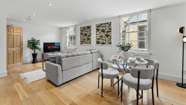Een moderne woonkamer met een grijze bank, een glazen eettafel met stoelen, een televisie en een potplant.