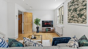 Een woonkamer met een houten vloer, een tv op een standaard, een potplant en een bank met kussens.
