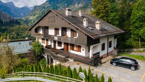 A two-story chalet with a balcony, set against a mountainous backdrop.
