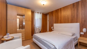A bedroom with a large bed, wooden walls, a wooden wardrobe, a small table with a vase, and a nightstand with a lamp.