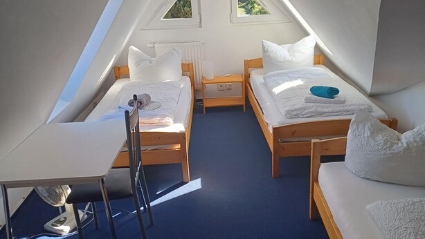 Une petite chambre mansardée de forme triangulaire, avec deux lits, une table et une chaise.