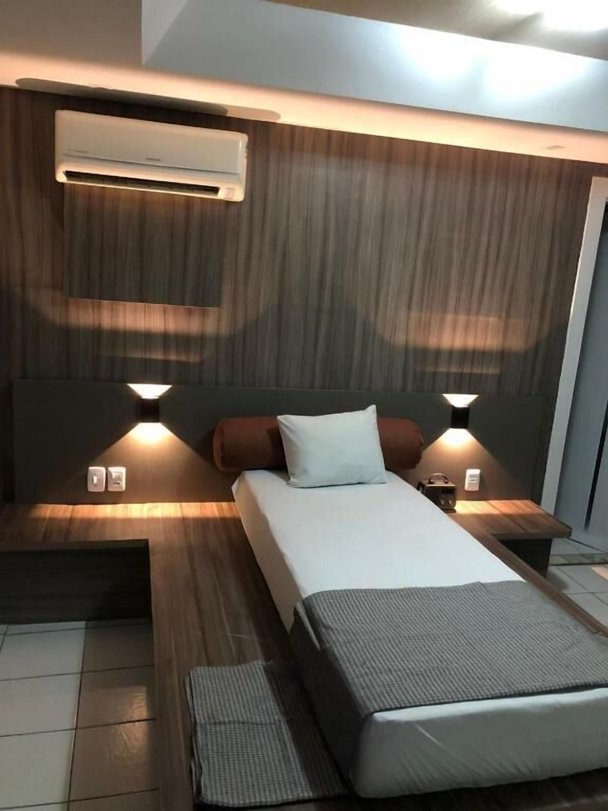 Una habitación de hotel moderna con una cama grande, dos mesitas de noche, aire acondicionado en la pared y una pequeña mesa con lámpara.