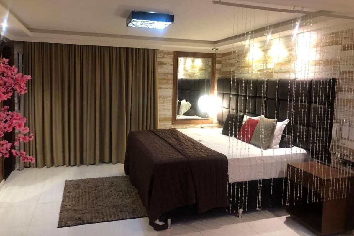 A modern bedroom with a large bed, a bedside table, a headboard with hanging lights, and a view of a cityscape through a window.