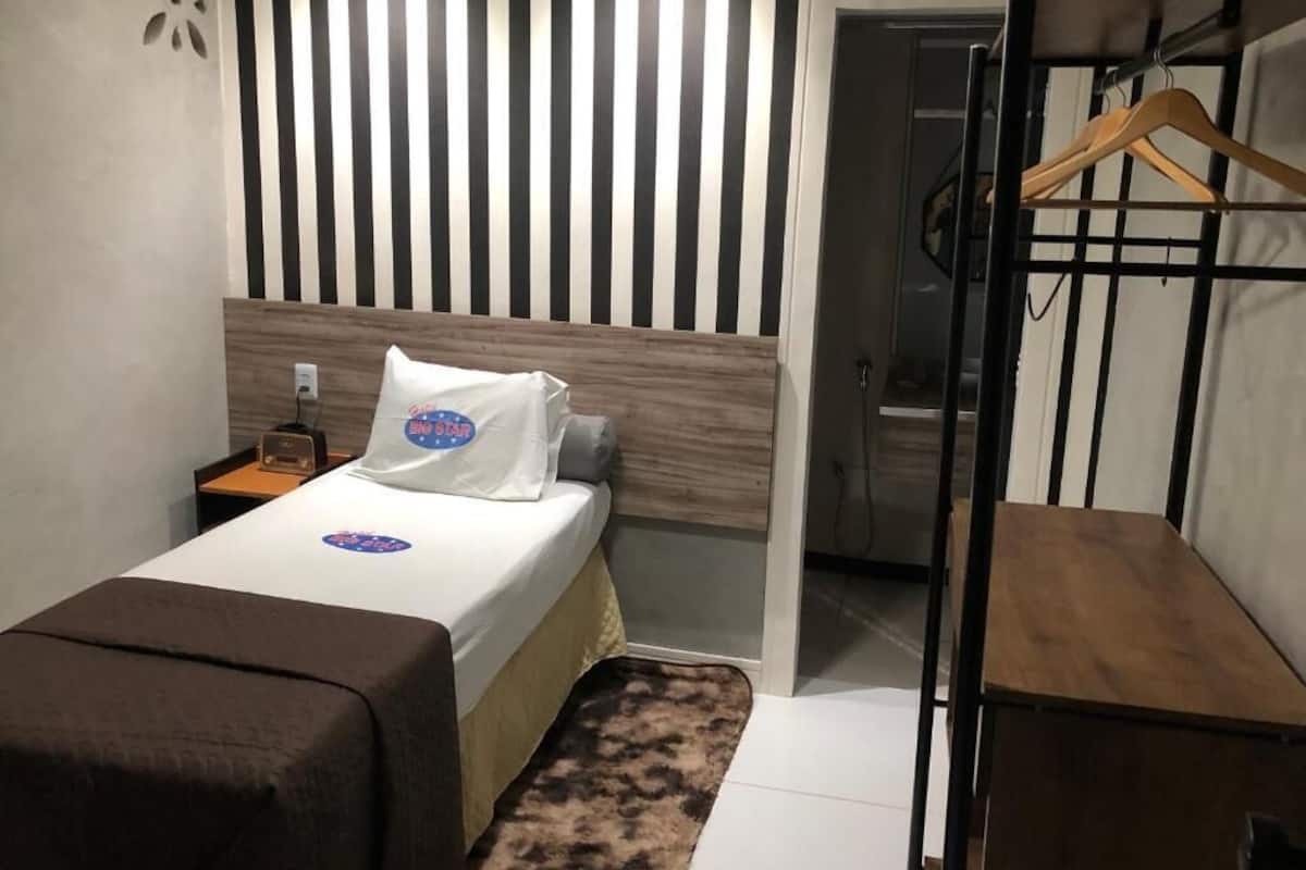 A compact hotel room with a bed, a bedside table, a hanging coat rack, and a small wooden bench.