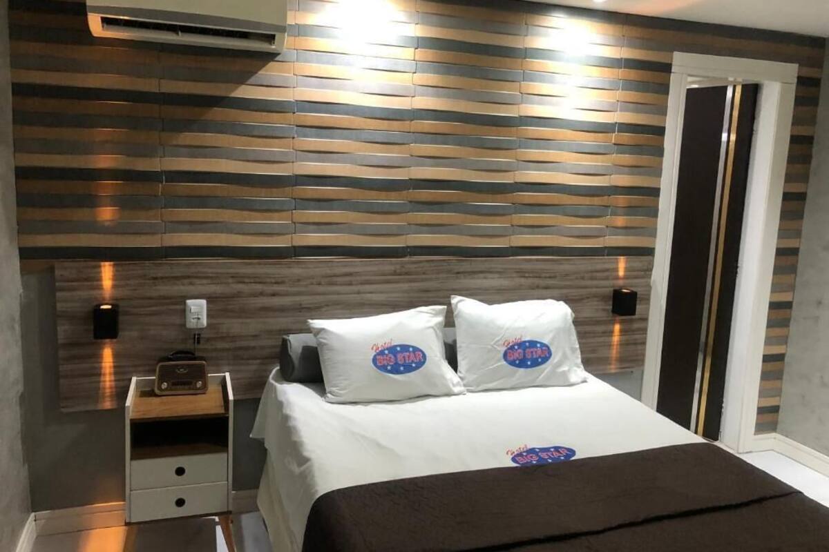 A modern bedroom with a wooden slat wall design, a bed with white bedding and two pillows, a nightstand with a radio, and a door with a golden handle.