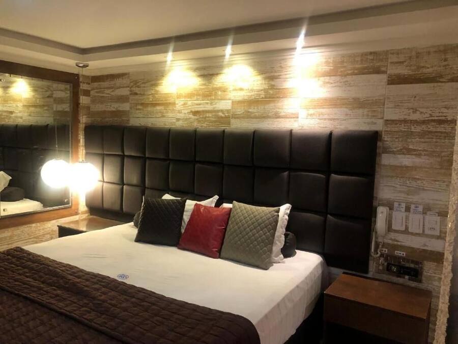 A modern hotel room with a large bed, a headboard with built-in lighting, a bedside table with a lamp, and a wall-mounted phone.