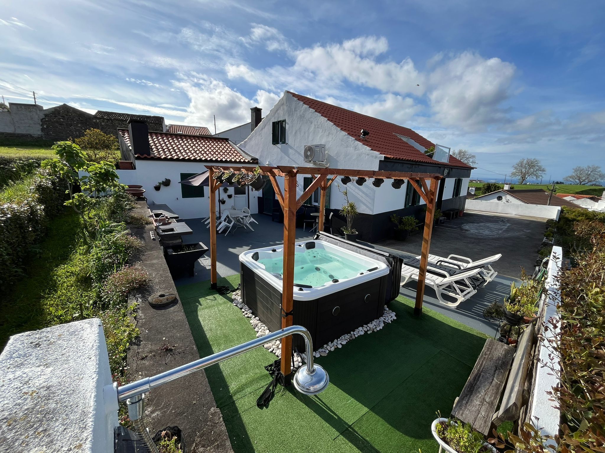 A hot tub under a wooden pergola in a backyard with a white house, green lawn, and a clear sky.