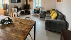 Un'ampia zona giorno con soffitto in legno, un divano grigio con cuscini gialli, un tavolo da pranzo in legno e un televisore montato a parete.