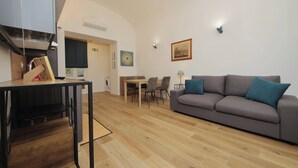 Ein modernes Wohnzimmer mit grauem Sofa, Holzboden, einem Esstisch mit Stühlen und einer Küchenzeile mit Schränken und einer Mikrowelle.