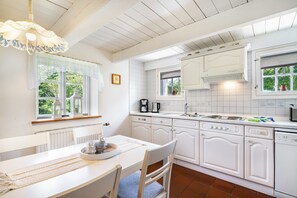 Une cuisine avec des meubles blancs, une table à manger avec des chaises, une fenêtre ornée de rideaux en dentelle et un luminaire suspendu.