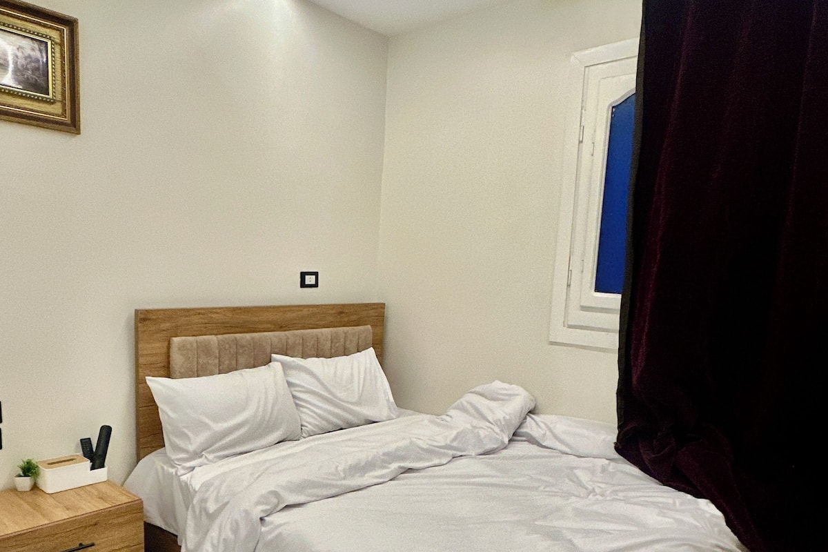 Une chambre avec un sol à carreaux, un cadre de lit en bois, du linge de lit blanc, une table de chevet avec une lampe et un tableau encadré au mur.