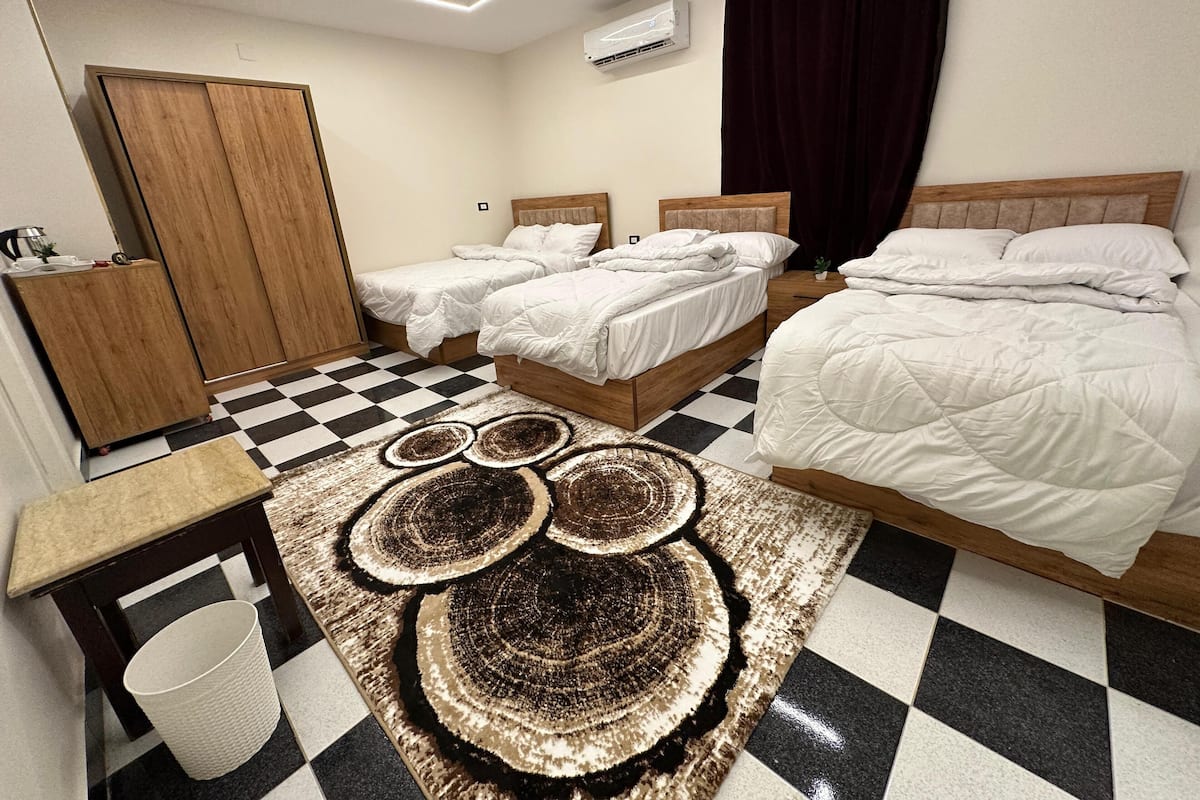 Une chambre d’hôtel avec deux lits simples et un lit double, un sol à carreaux, une armoire en bois, une petite table avec une lampe et un tapis décoratif.