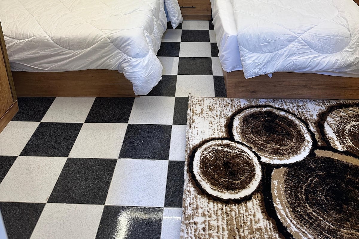 Une chambre d’hôtel avec deux lits, un sol à carreaux, un climatiseur, une table de chevet avec une télécommande et un rideau foncé.