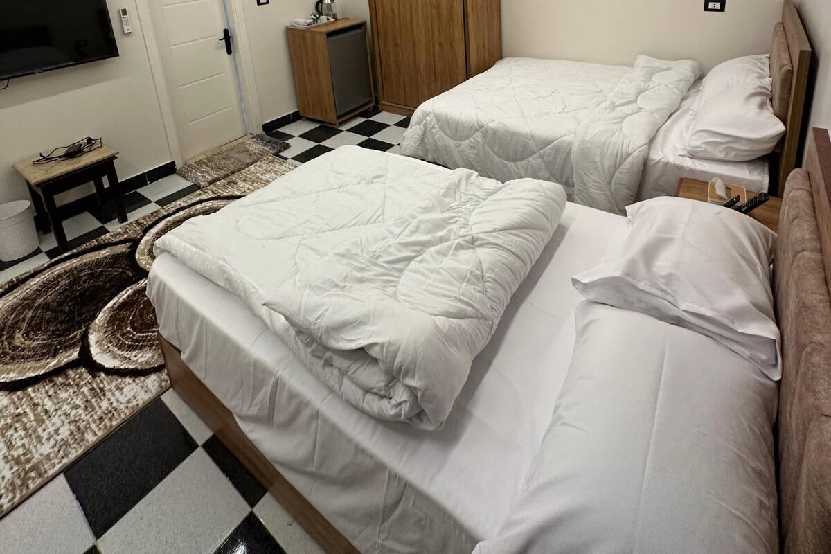 Une chambre d’hôtel moderne avec deux lits, un téléviseur à écran plat fixé au mur, une armoire et un sol à carreaux.
