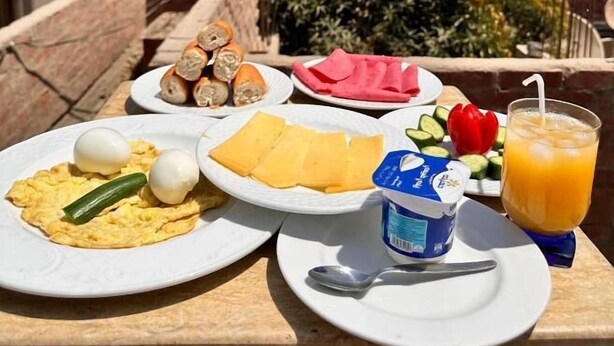 A table with breakfast items including eggs, cheese, and a drink, on a balcony with buildings and greenery in the background. - Great Pyramids Lodge (Giza)